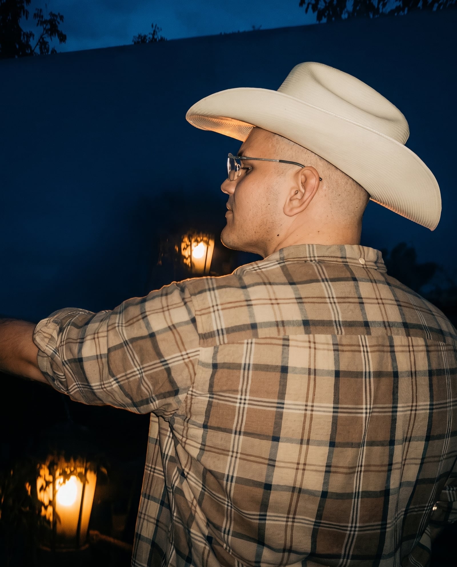 Dillien Gutknecht im Profil, mit hellem Cowboyhut und Brille, abends vor einer blauen Wand, links zwei warm leuchtende Laternen.